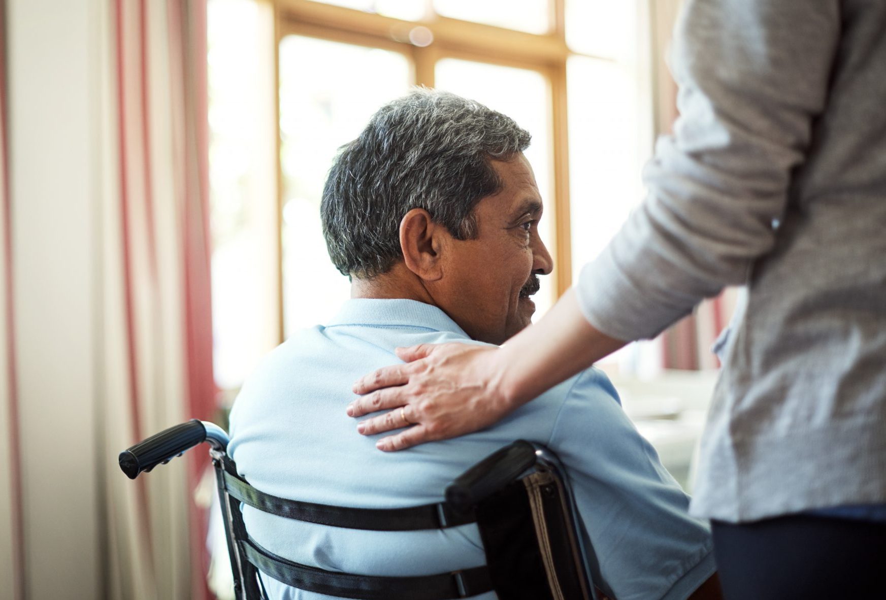 Un homme d’âge moyen à peau brune assis dans un fauteuil roulant se faisant réconforter par une autre personne dont la main est posée sur son dos.